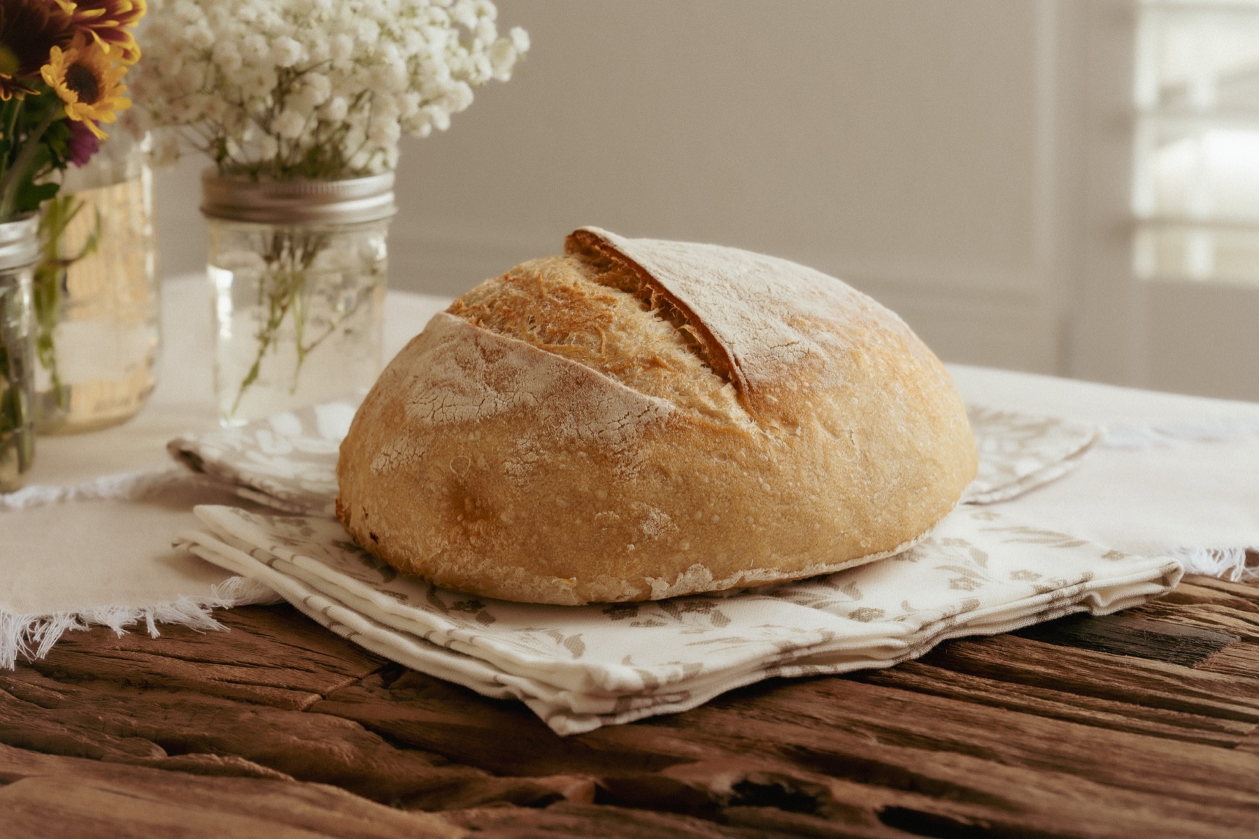 A round loaf of bread on folded cloth beside flowers.