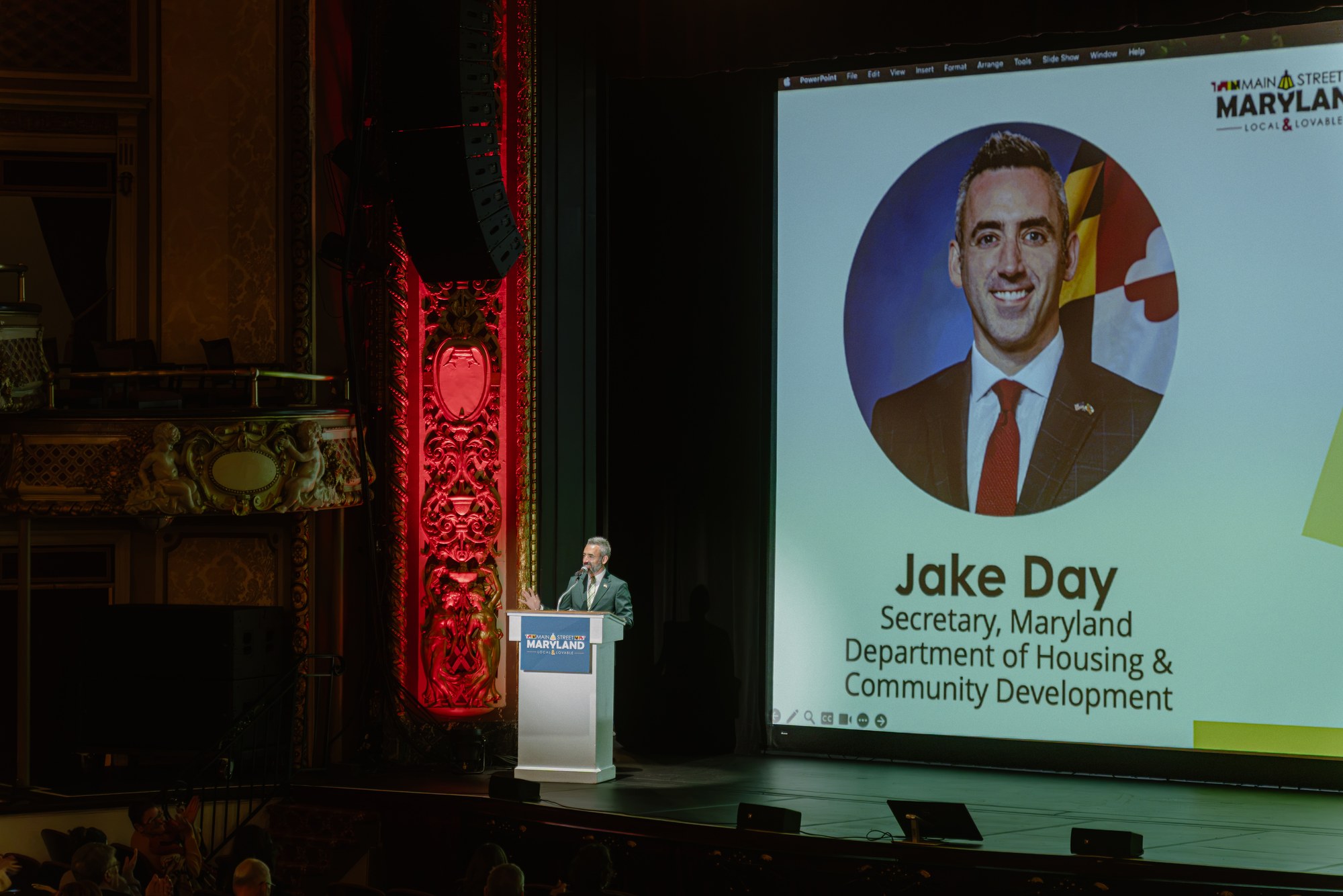 A speaker on the Maryland Theatre stage alongside the conference presentation screen